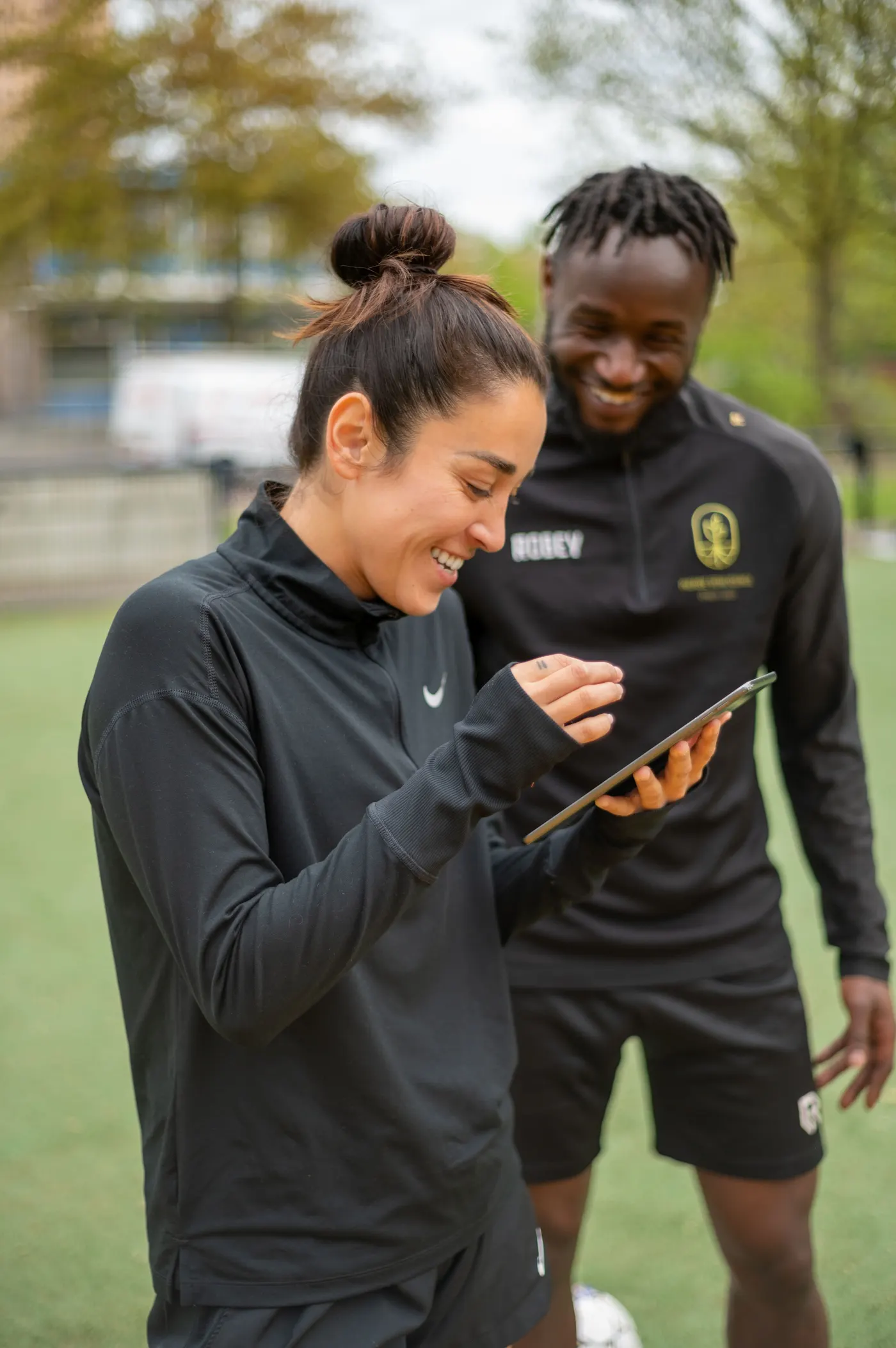 Twee coaches van Voetbal Dream Coach bekijken lachend een tablet langs het veld